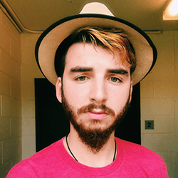 A young man with a beard and mustache, wearing a tan hat and a pink shirt, poses for a selfie in a well-lit indoor setting.