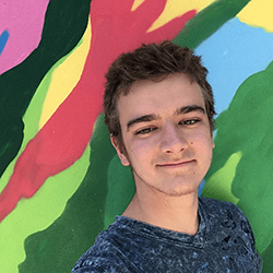 A young man with short, light brown hair smiles at the camera while standing in front of a colorful, abstract mural featuring vibrant shades of green, pink, and yellow. He wears a blue patterned shirt.