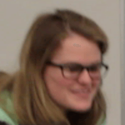 A slightly blurred image of a person with long, wavy hair, wearing glasses and a light-colored hoodie, smiling or laughing while facing slightly to the side.