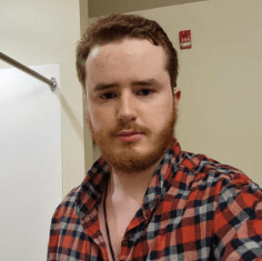 A young man with a beard wearing a red and black plaid shirt, standing in a room with a whiteboard in the background.