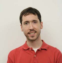 A man with short dark hair smiles while wearing a red polo shirt against a plain white background.