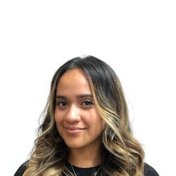 A young woman with long, wavy hair that is dark at the roots and lighter towards the ends, smiling softly against a plain white background. She is wearing a black t-shirt and has a few delicate necklaces.
