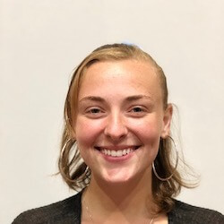 A smiling young woman with shoulder-length, wavy blonde hair, wearing hoop earrings and a dark cardigan, poses against a plain light background.