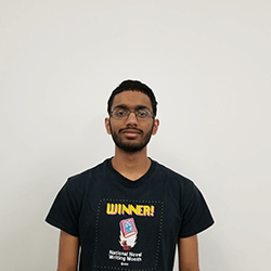 A young man with glasses stands against a plain white background, wearing a black t-shirt that features the word 