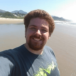 A person with curly hair and a beard is smiling at the camera while standing on a beach. The ocean and mountains are visible in the background, and the weather appears sunny.