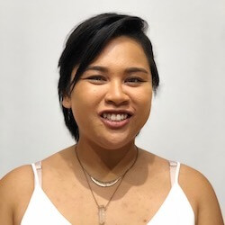 A smiling person with short, dark hair wearing a white top and layered necklaces, standing against a light background.