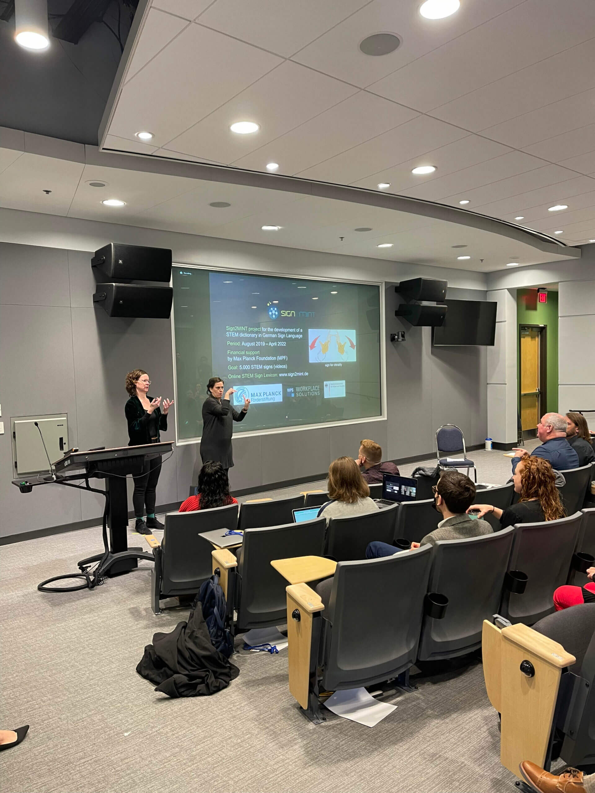 A presentation is taking place in a lecture hall, with two speakers gesturing towards a large screen displaying text and images. An audience is seated in rows, focusing on the presentation. The room is equipped with audio-visual equipment.