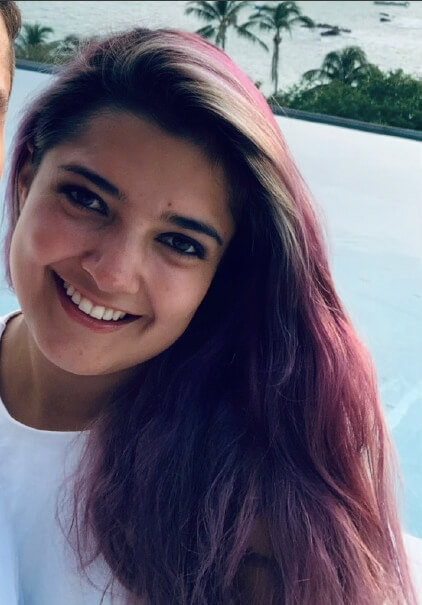 A smiling young woman with long, wavy hair that has pink highlights, wearing a white top. The background features a blurred outdoor setting, suggesting a tropical location.