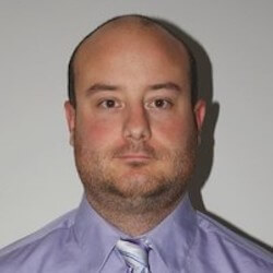 A man with a bald head and a short beard is wearing a light purple dress shirt and a patterned tie. He is facing the camera with a neutral expression against a plain background.