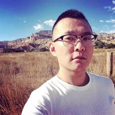 A person with short hair and glasses stands in a grassy field, with rocky hills and a blue sky in the background.