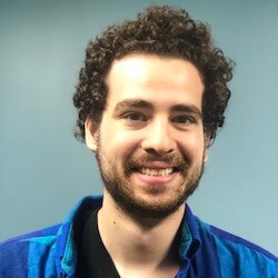A smiling individual with curly hair is wearing a blue and black checkered shirt. The background is a solid light blue color.