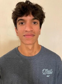 A young man with short, wavy hair is facing the camera, wearing a gray O'Neill t-shirt. He has a neutral expression and is positioned against a light-colored wall.