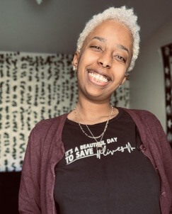 A smiling individual with short, curly blonde hair is wearing a black t-shirt that reads 