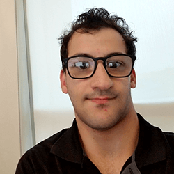 A young man with curly hair and glasses, wearing a black shirt, is smiling at the camera. The background appears to be a light-colored wall with natural light coming through.
