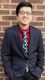 A young man stands confidently in front of a brick wall, wearing a dark suit jacket over a red shirt and a brightly patterned tie. He has glasses and is smiling, with his hands in his pockets.