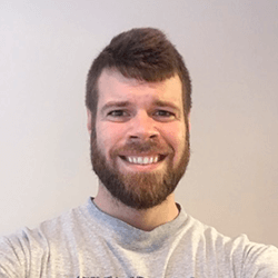 A smiling man with a short beard and tousled hair, wearing a gray t-shirt, poses for a selfie against a light background.
