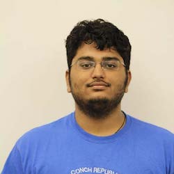 A young man with curly black hair and glasses, wearing a blue t-shirt, is standing against a plain light background.