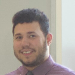 A man with short, dark hair and a beard smiling while wearing a purple dress shirt and tie, standing in front of a light background.