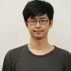 A young man with short black hair and glasses, wearing a gray t-shirt, stands against a plain light background, smiling softly at the camera.