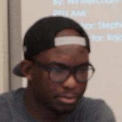 A young man wearing a black cap and glasses is sitting in front of a presentation screen. He appears focused, with a slight frown, as he listens or observes something.