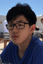 A young person with short black hair and glasses, wearing a blue shirt, is sitting outdoors with a neutral expression. The background shows a sandy area and some people in casual clothing.