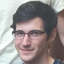 A young man with dark, short hair and glasses smiles at the camera, with a blurred background of other people.
