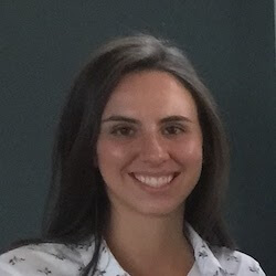 A young woman with long, dark hair smiles at the camera, wearing a white shirt with a subtle pattern. The background is dark, suggesting an indoor setting.