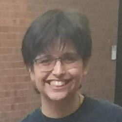 A person with short dark hair and glasses smiling in a casual setting with a brick wall in the background.