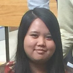 A young woman with long black hair is smiling softly while sitting in a casual indoor setting. She is wearing a patterned top and is partially surrounded by people in the background.