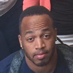 A close-up of a man with short hair and a light beard, wearing a navy blue shirt. He has a neutral expression and is looking directly at the camera.