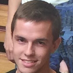 A young man with short brown hair is smiling at the camera, seated in a casual setting.