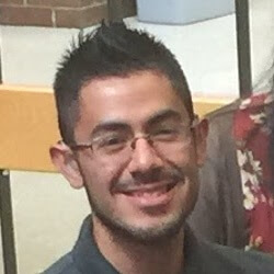 A smiling young man with short, spiky hair and glasses, wearing a dark-colored shirt, is seated in a casual environment with a wooden bench in the background.