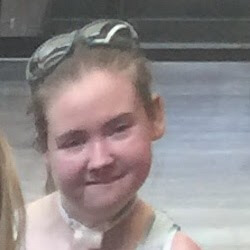 A young person with light brown hair, wearing sunglasses, smiling slightly, in a casual setting with a blurred background.