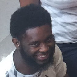 A man with a short, curly black beard and hair is smiling. He is wearing a white shirt with black stripes and is seated, with another person partially visible in the background.