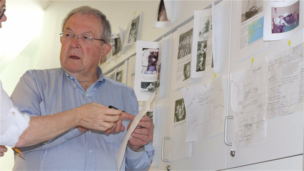 A man in a light blue shirt is engaged in a discussion with another person, holding a piece of paper and looking at a wall covered with various printed images and documents.