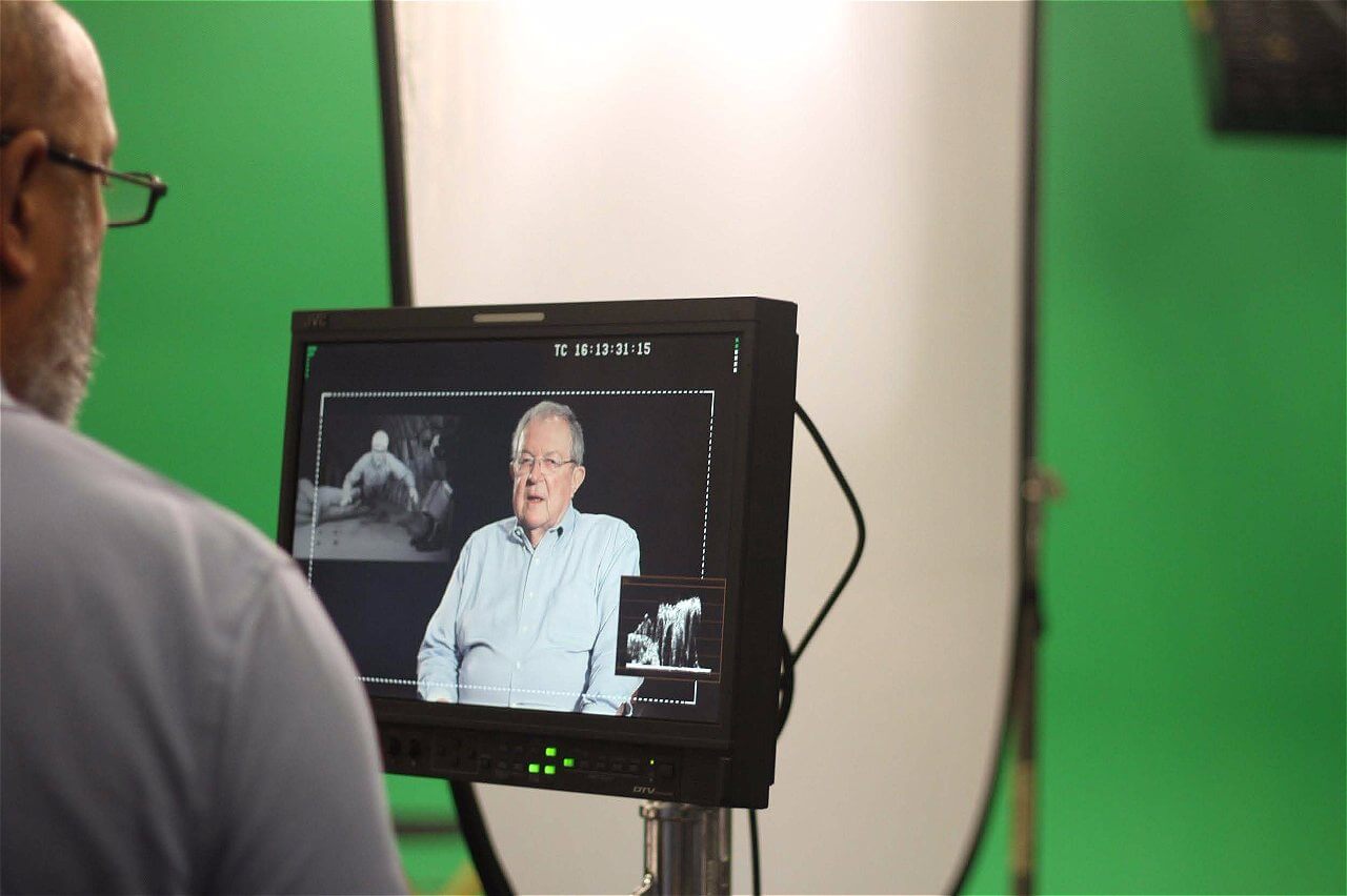A person is seen from behind, looking at a monitor that displays an interview with an older man sitting in front of a green screen. The monitor shows the man speaking, with another small image in the corner. Soft lighting is set up in the background.