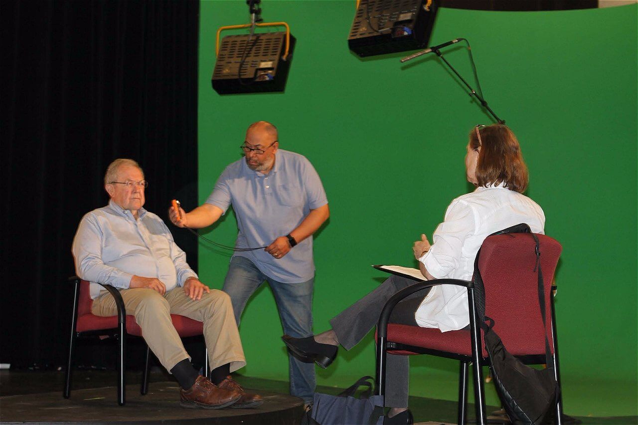 A man in a blue shirt stands beside an older man seated in a light blue shirt, while a woman in a white jacket sits in a chair, taking notes. The background is a green screen, and there are studio lights overhead.