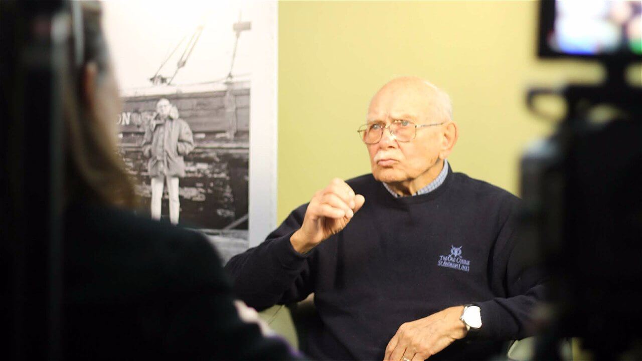 An elderly man with glasses and a mustache sits in front of a camera, gesturing while speaking. He is wearing a dark sweater and is positioned against a background featuring a black-and-white photograph. The image captures a moment of an interview or discussion.