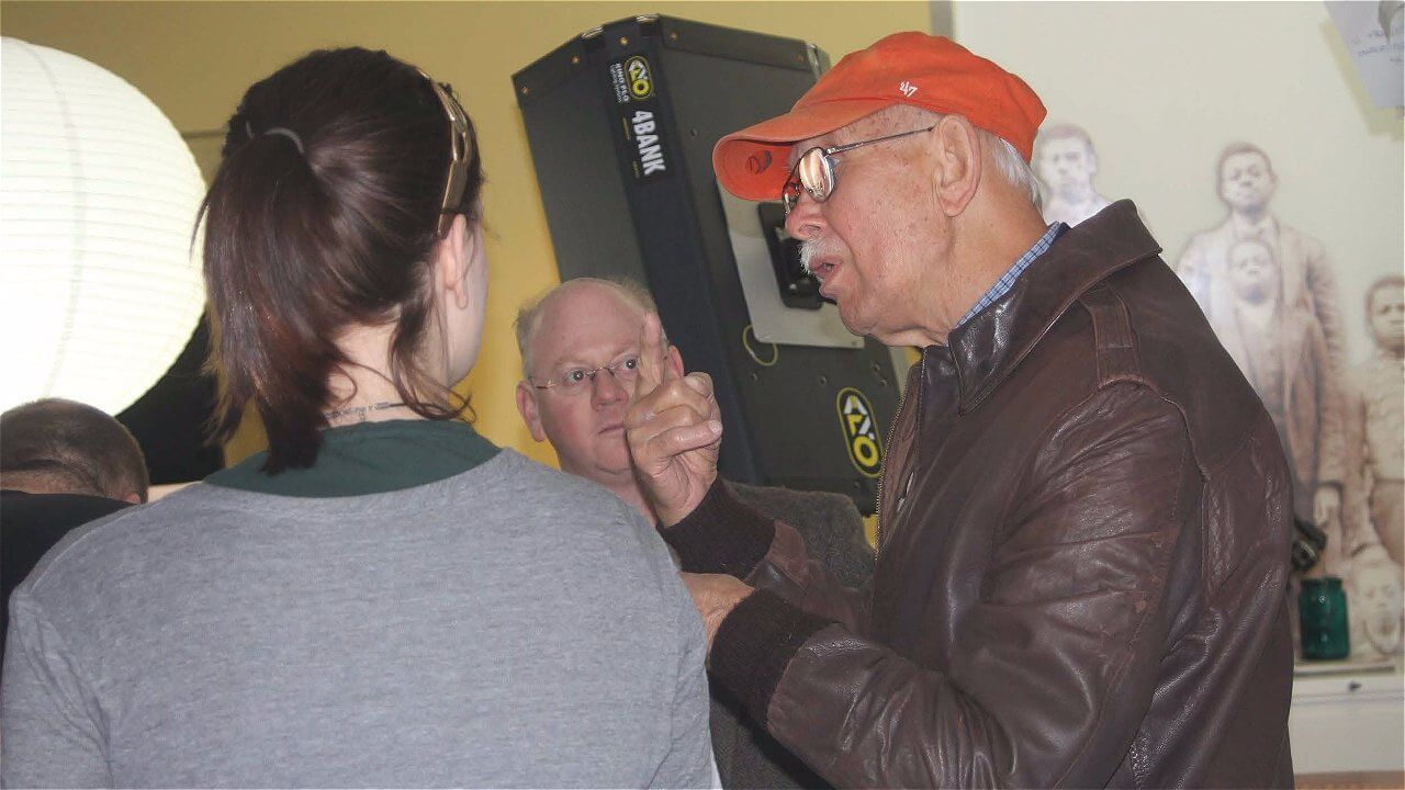 A group of three people engaged in conversation indoors. One person, wearing an orange cap and a brown leather jacket, is animatedly speaking, while another person, with glasses, listens attentively. A third person, with their back to the camera, is also part of the discussion. The background features soft lighting and wall decor.