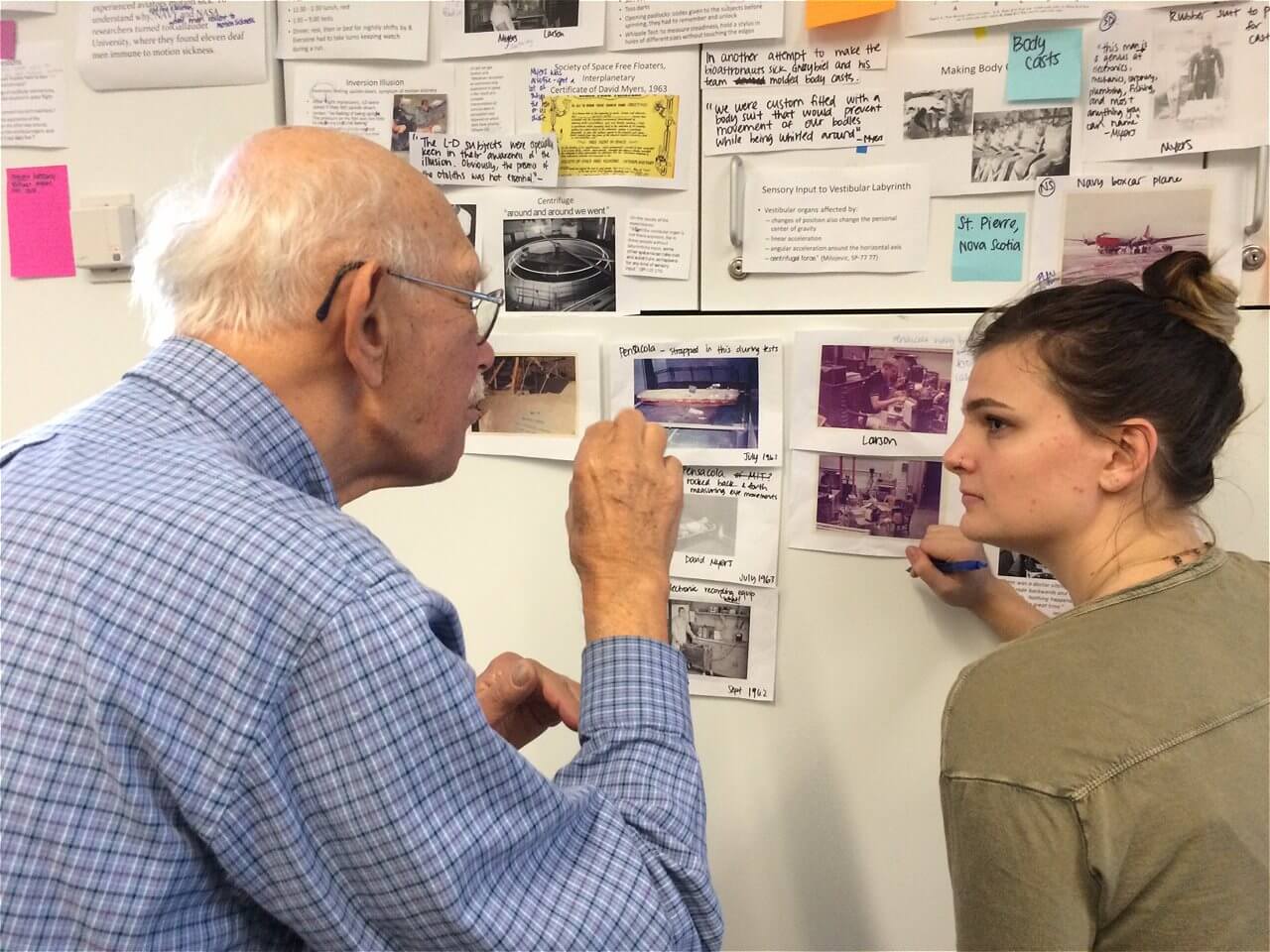 An elderly man with glasses and a blue checkered shirt is engaged in conversation with a young woman in a casual green shirt, both looking at a wall covered with various photographs and notes. The man is gesturing with his hand as he speaks, while the woman appears to be listening attentively.