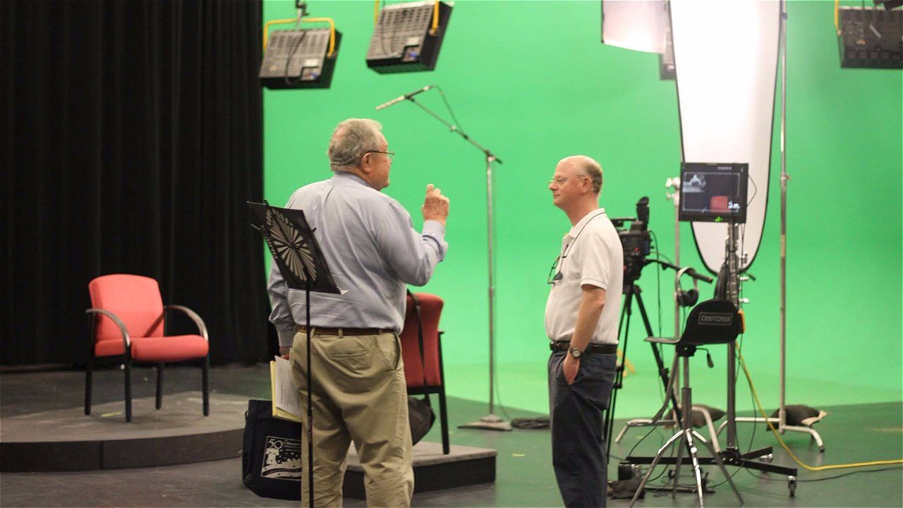 Two men are engaged in conversation on a studio set with a green screen backdrop. One man is gesturing with his hand while the other listens. A red chair is positioned nearby, and there are several lighting equipment and microphones set up around them.