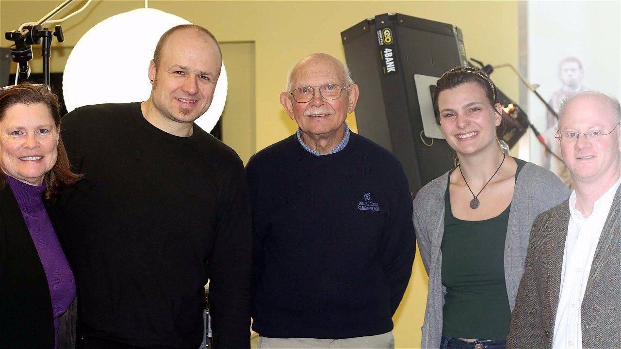 A group of five people stands together, smiling for the camera. They are positioned in a well-lit room, with studio equipment in the background. The individuals include two men and three women, each wearing casual clothing.