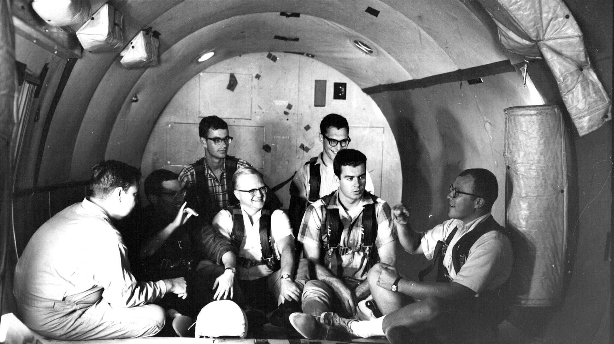 A group of six men seated inside a cylindrical space resembling a spacecraft or experimental environment, engaged in discussion. They are dressed in casual clothing and appear to be working together, with various equipment and materials scattered around them. The interior has a metallic appearance with overhead lights and fabric drapery.