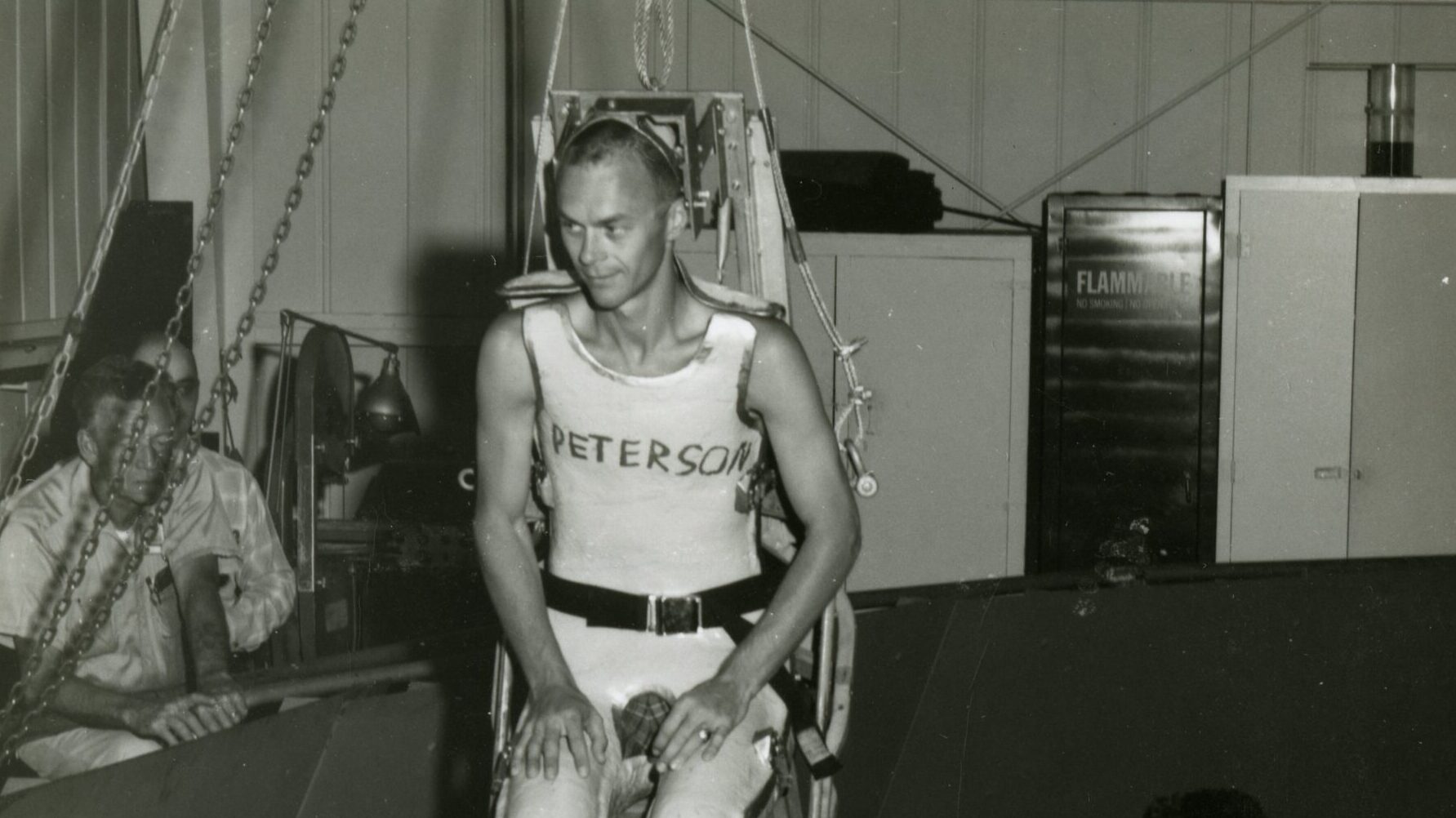 A black and white photograph of a man seated in a harness apparatus, preparing for a test or experiment. He is wearing a sleeveless shirt with "PETERSON" printed on it. Two other individuals are present in the background, one of whom appears to be observing or adjusting equipment. The setting looks like a laboratory or testing facility.
