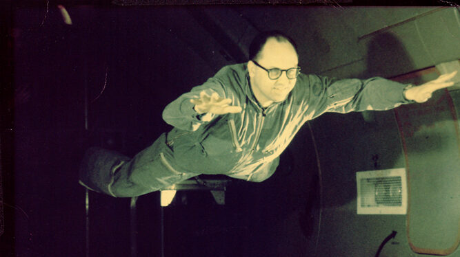 A man wearing glasses and a jumpsuit is floating in a zero-gravity environment, with his arms outstretched as if flying. The background appears to be the interior of an aircraft or a similar facility designed for testing.