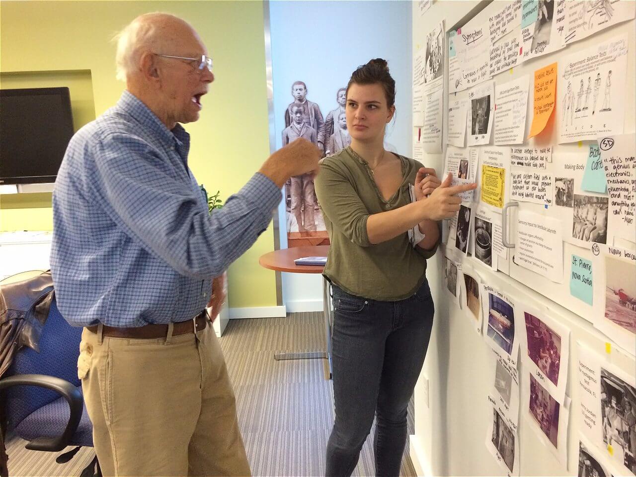 An older man with glasses is animatedly speaking while gesturing with his hands, and a young woman is listening attentively, standing beside him and pointing at a wall covered with various notes, images, and documents. The setting appears to be an office or collaborative workspace.