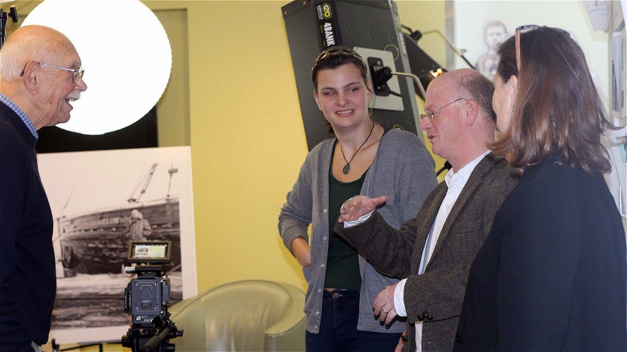 A group of four people engaged in conversation in an indoor setting, with a camera and photography equipment nearby. One person is older, wearing a dark sweater, while the others are younger, dressed casually. Behind them, there is a large circular light and a black-and-white photograph of a ship on the wall.