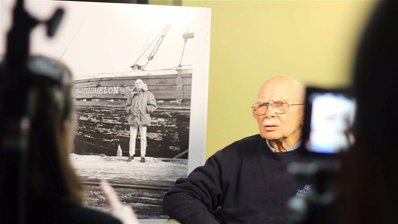 An elderly man sits in front of a large black-and-white photograph of a younger person standing next to a boat. The man appears to be engaged in conversation, looking thoughtfully at the camera. In the foreground, part of a person's hand is visible, gesturing towards him, suggesting an interview or discussion is taking place.