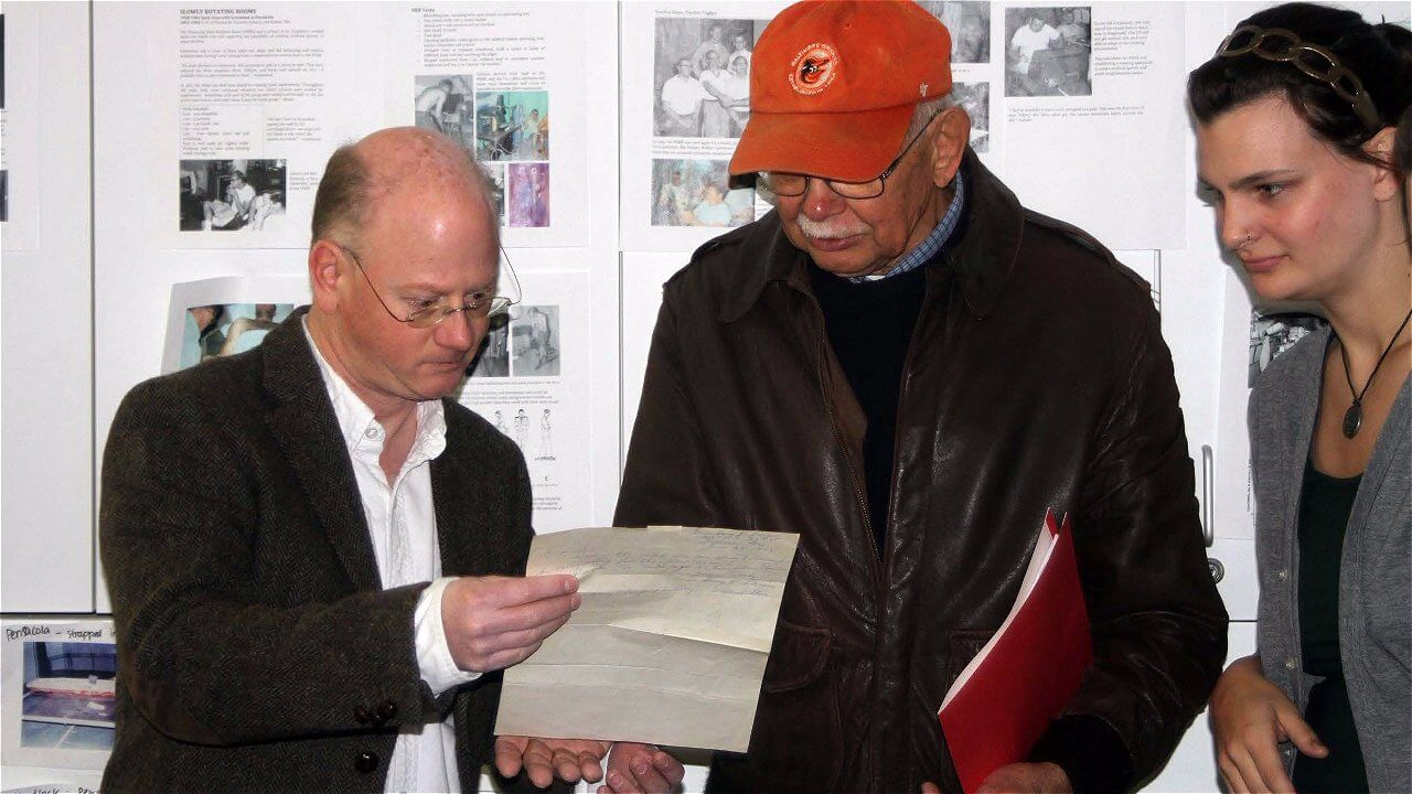 A group of three people is gathered around a piece of paper. One man, wearing a dark jacket and glasses, is pointing to the paper while another older man, wearing an orange baseball cap and a brown jacket, is looking closely at it. A woman, dressed in a dark top, stands beside them, observing the interaction. In the background, there are various documents and photographs displayed on a wall.