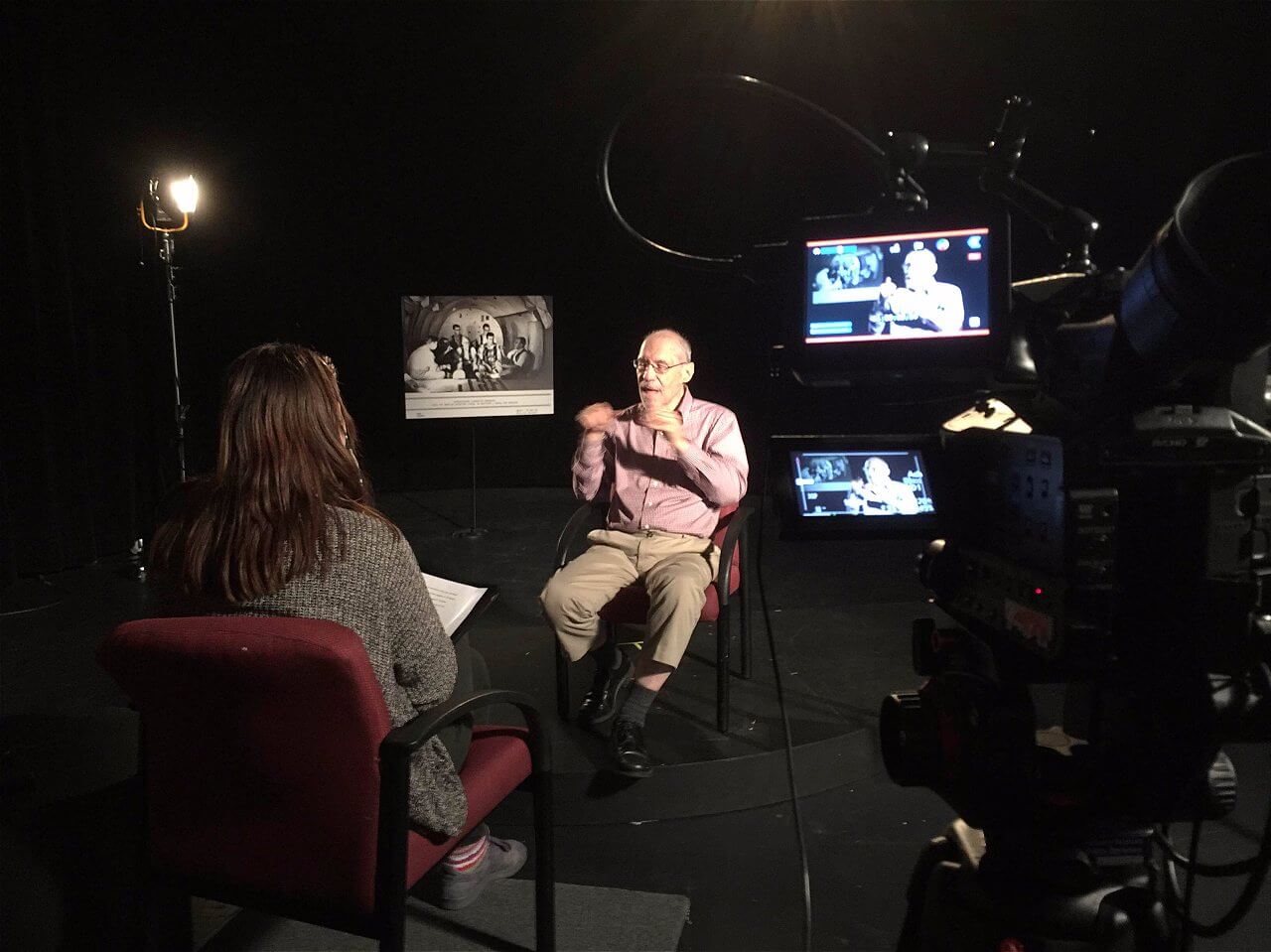 A man in a pink shirt sits in a chair during an interview, gesturing while speaking. He is facing a woman in a gray sweater who is seated across from him, holding a notepad. The setting is dimly lit with a spotlight on the man, and there are cameras positioned to capture the conversation. In the background, a black-and-white photograph is visible on the wall.